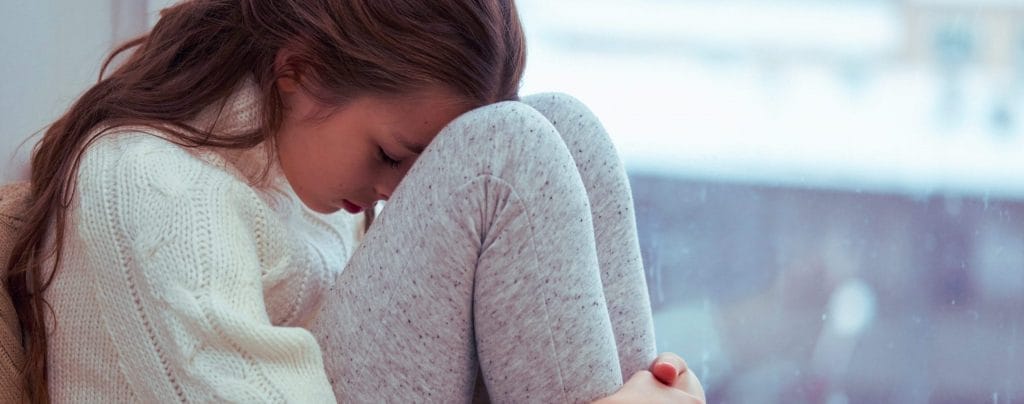 Young girl sitting near a window holding her head to her knees