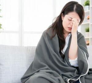 Woman with dark hair sitting on a couch with a gray blanket wrapped around her shoulders holding the center of her forehead, seemingly in pain