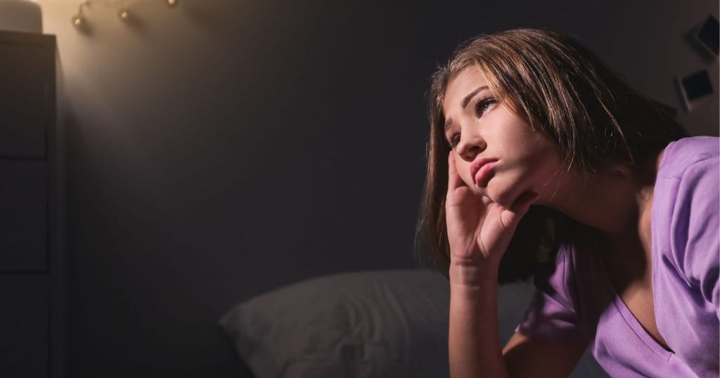 Girl wearing purple shirt sitting in a dark room with sad look on her face