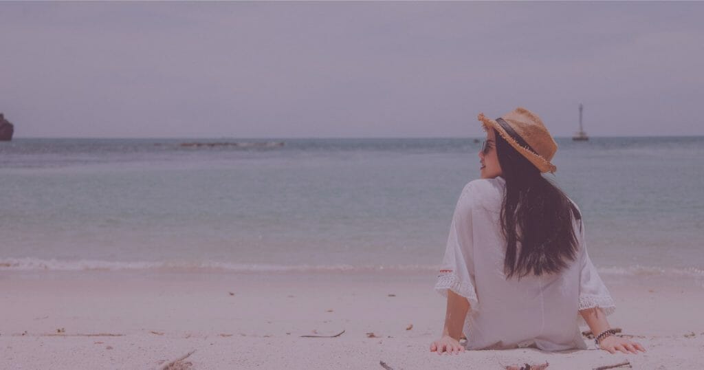 Woman with dark hair wearing a white shirt, sunglasses and straw hat sitting on the beach looking out at the water