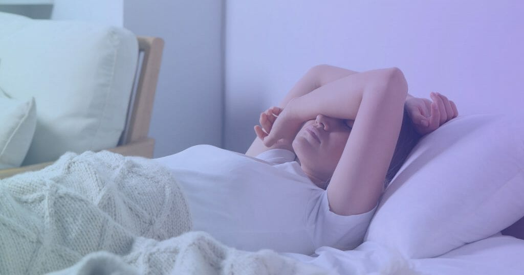 Woman wearing white shirt laying in bed holding her arm over her eyes to block light