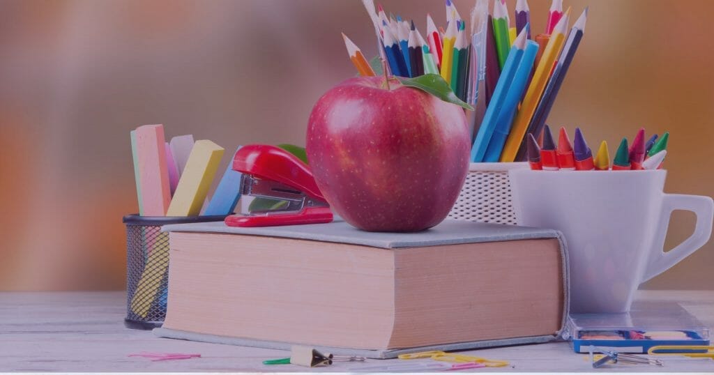 A red apple sitting on a book, colored pencils and school supplies