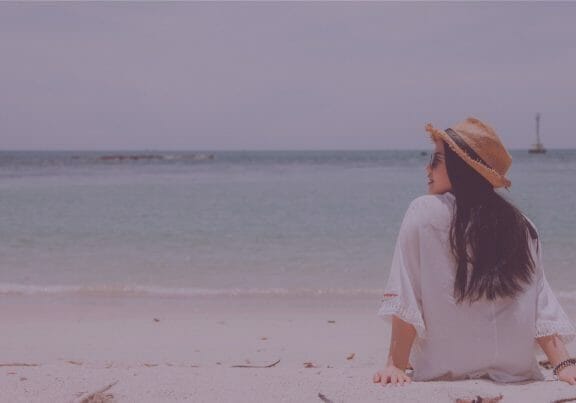 Woman with dark hair wearing a white shirt, sunglasses and straw hat sitting on the beach looking out at the water