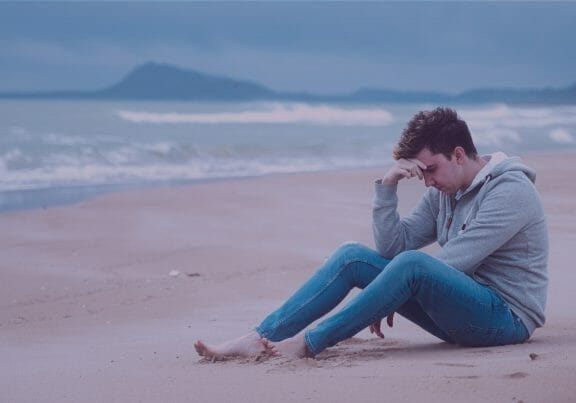 Man wearing jeans and a gray sweatshirt sitting on a deserted beach holding his head