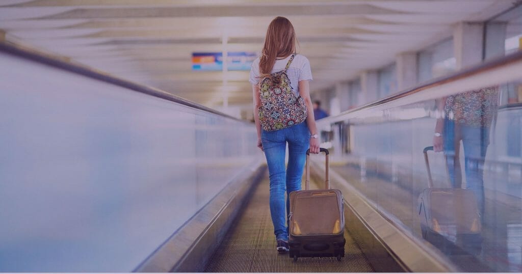 Woman wearing blue jeans and white top walking in airport wearing a backpack and carrying a roller suitcase