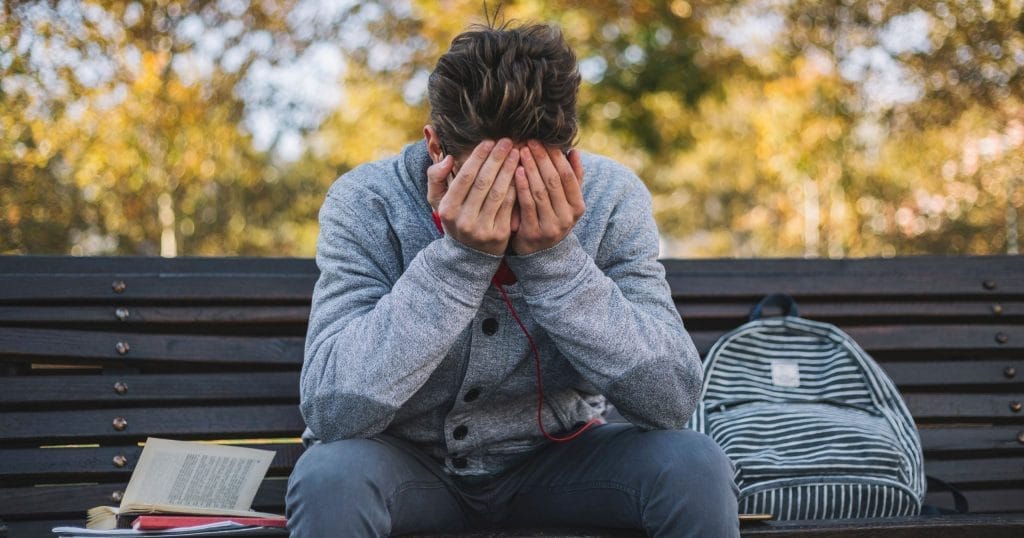 College age young man sitting on bench outside with books next to him holding his face and head in pain