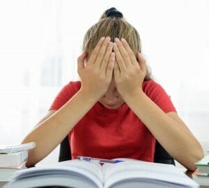 Girl with blonde hair in pony tail wearing a red shirt. She's sitting at a desk with a book on it and holding her head in her hands as if she is in pain.