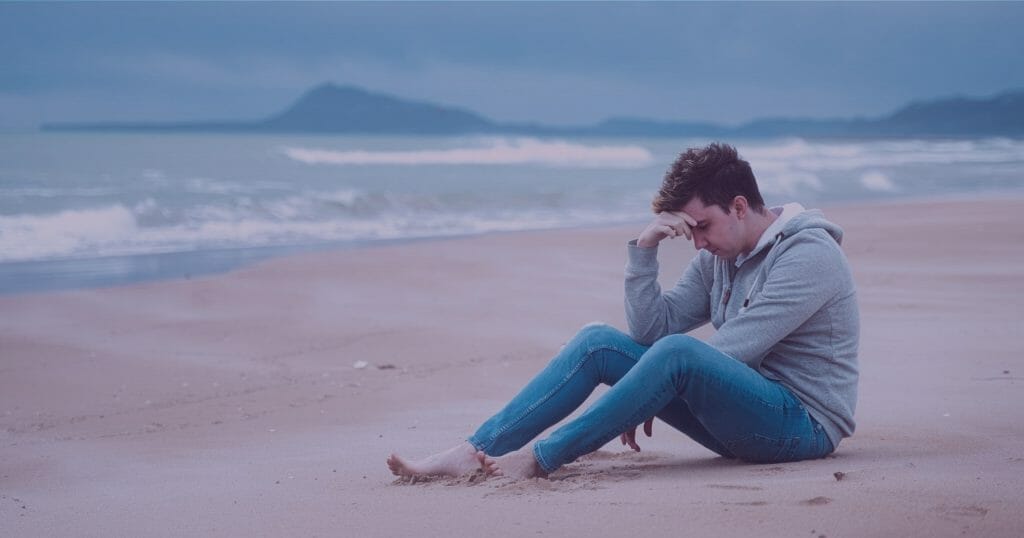 Man wearing jeans and a gray sweatshirt sitting on a deserted beach holding his head