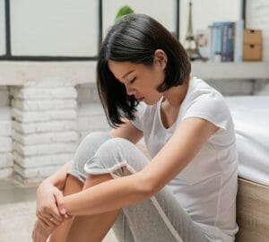 Woman with dark hair wearing a white tshirt and gray sweatpants sitting on the floor at the foot of a bed with her arms wrapped around her knees feeling lonely