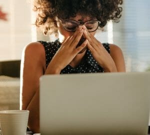 Woman wearing glasses sitting at a computer holding her eyes and head in pain