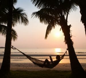 Woman laying in hammock hung between two palm trees on a beach at sunset reading a book