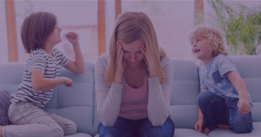 Woman with blonde hair sitting on sofa holding her head with her hands while two young blonde children jump next to her