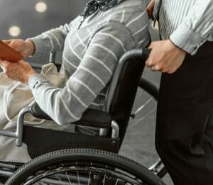 Older woman with white hair being pushed in wheelchair in an airport by a man wearing a button down shirt and pants